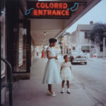 A photograph of a black mother and her girl standing below a neon sign that says 'colored entrance'