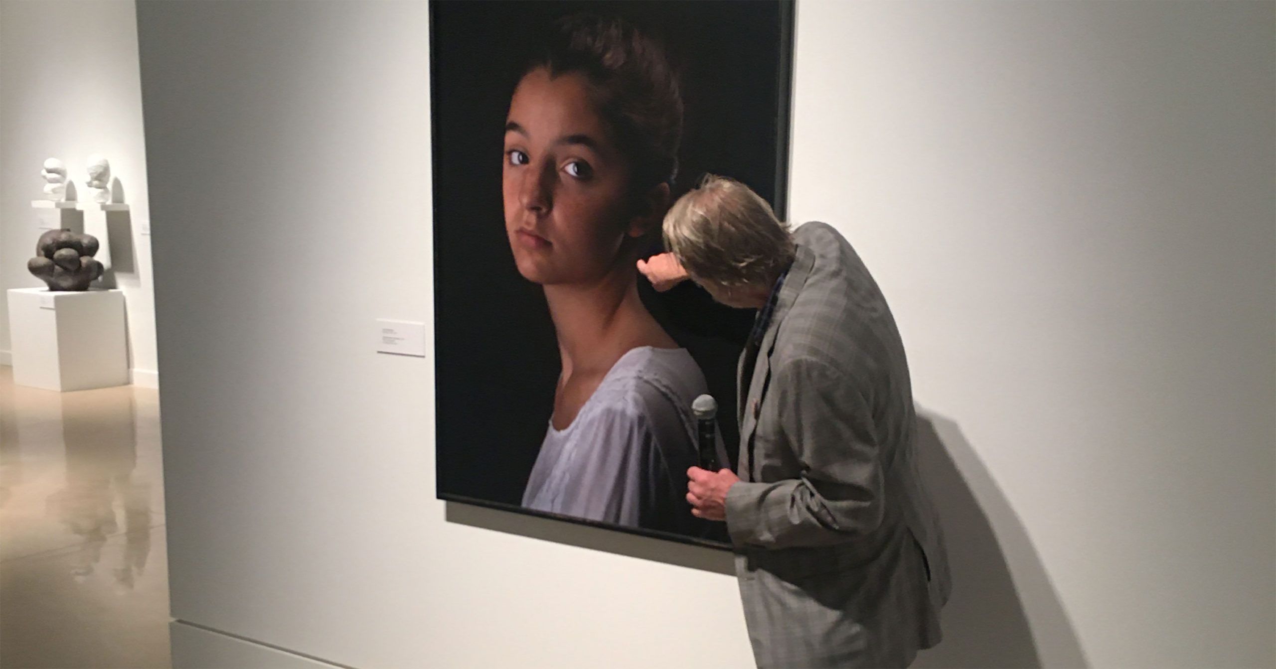 a man with a microphone points at a detail on a painting on the gallery wall