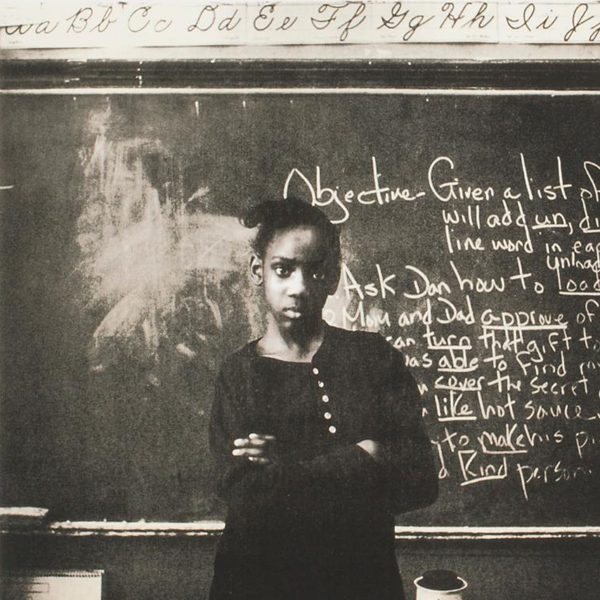 A young Black woman stands arms folded in front of a chalk board looking at the viewer