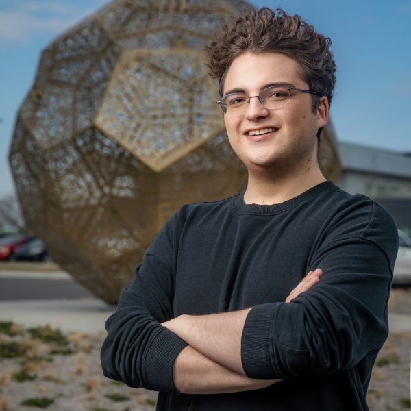 Austin Storie stands in front of a large, brass, spherical sculpture.