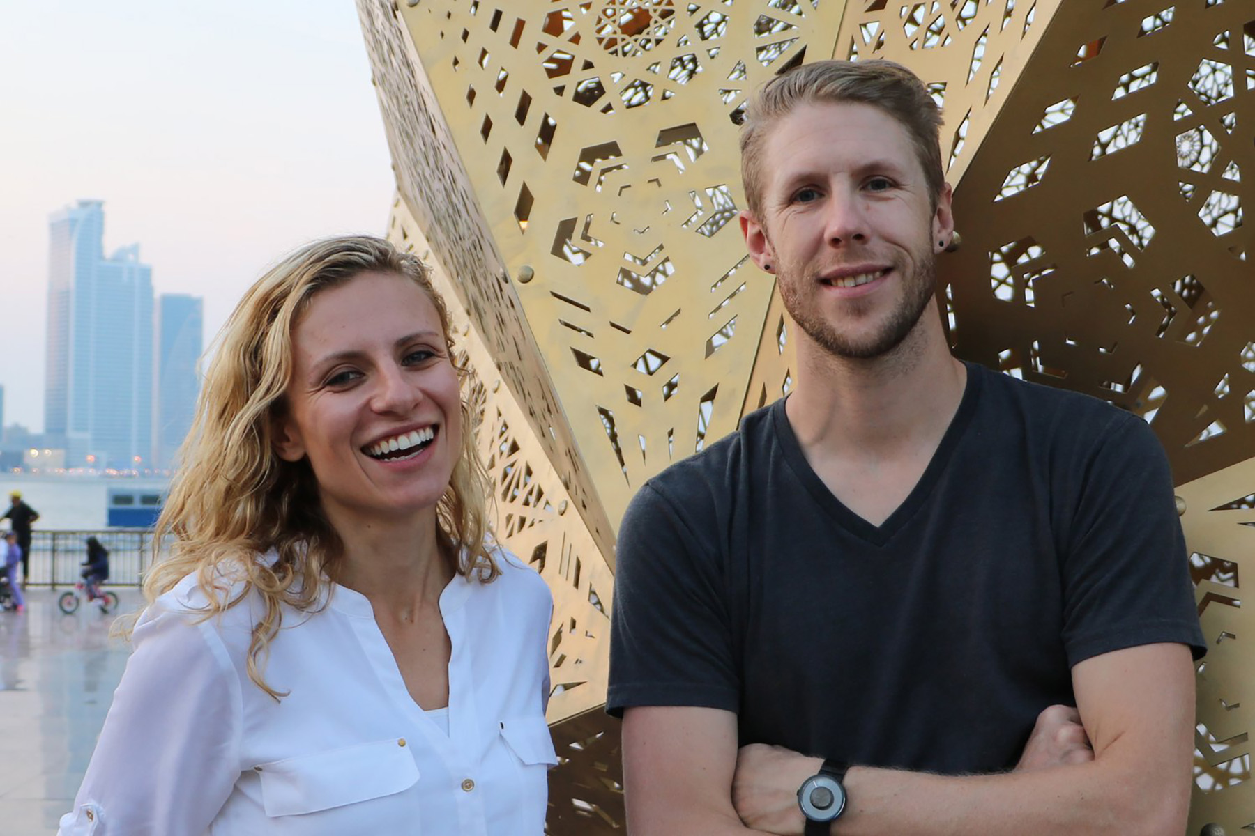 Two blond artists, Yelena Filipchuk and Serge Beaulieu are standing in front of a lacy, brass sculpture they created.