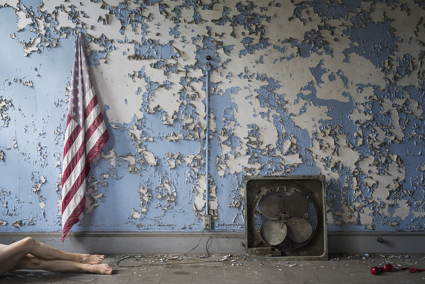 a wall with peeling blue paint, an American flag, a box flag and a red phone. From the left, a pair of female legs emerge.