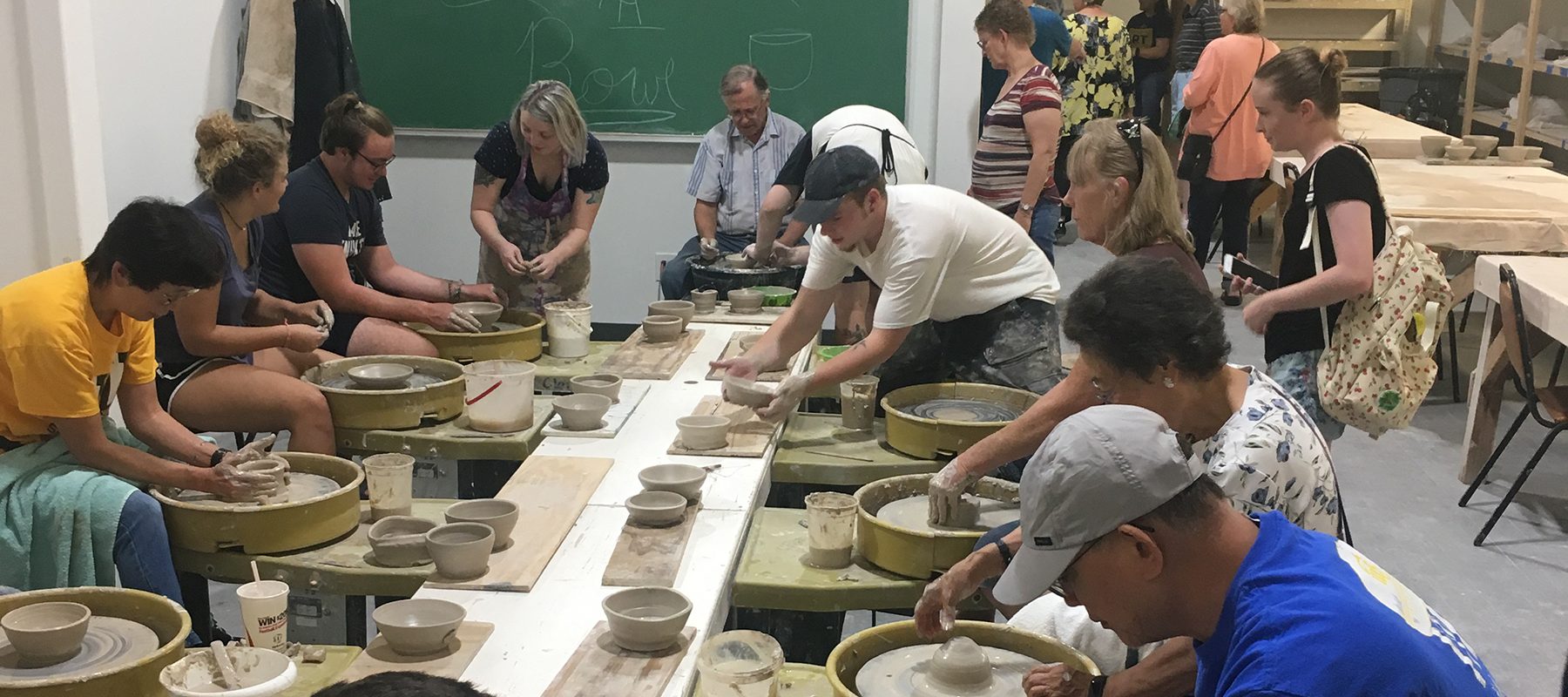 Eleven people sit around a table at potter's wheels, creating bowls from clay.