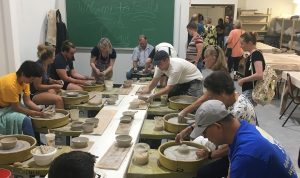 Eleven people sit around a table at potter's wheels, creating bowls from clay.