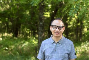 A man with glasses stands in front of a wooded area.