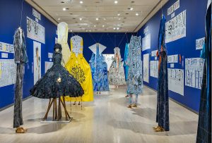 Seven sculptures shaped like dresses are positioned in a blue room.