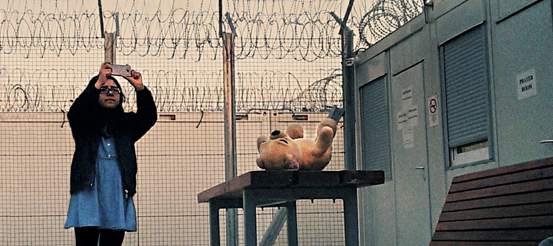 A girl is taking photos inside if a fence topped with razor wire. A teddy bear sits on a table nearby.
