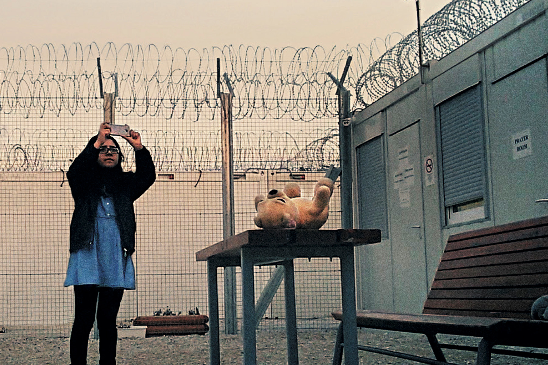 A girl is taking photos inside if a fence topped with razor wire. A teddy bear sits on a table nearby.