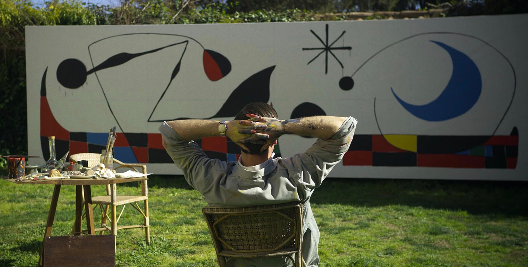 In a still from the film Miró, the actor playing the artist sits with his back to us as he looks at a large painting he is working on.