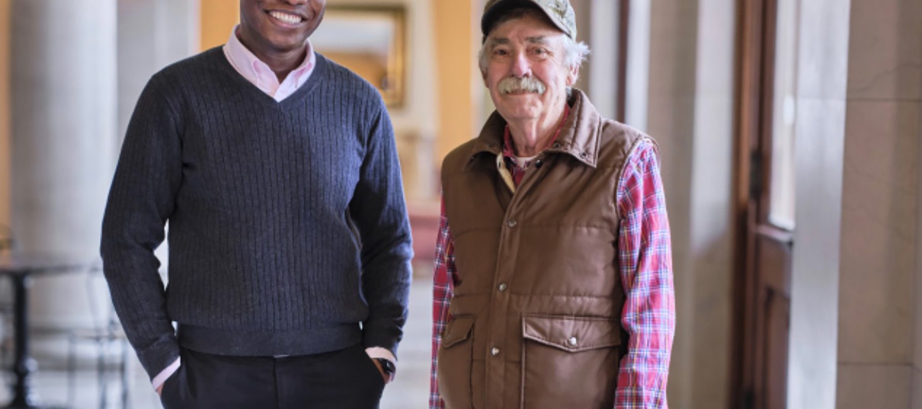 A middle-aged black man and a senior white man stand together in a hall with columns.