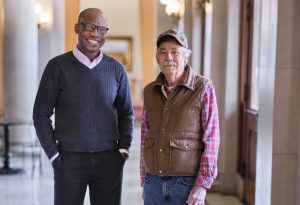 A middle-aged black man and a senior white man stand together in a hall with columns.