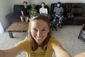 Four people sit on a couch in the background. In the foreground, a smiling woman facing the camera takes their photo.