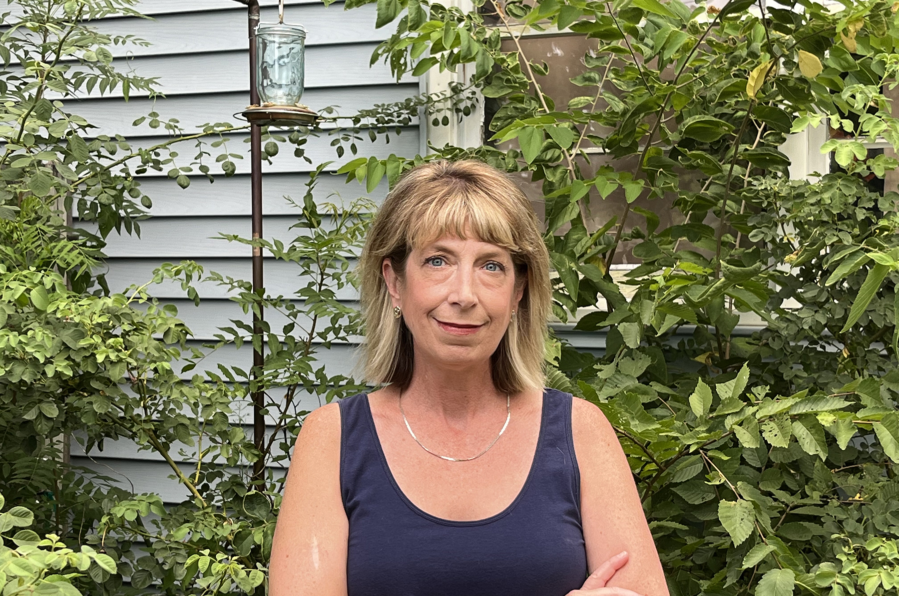 A blonde woman stands next to a bird feeder in front of some foliage by a house.