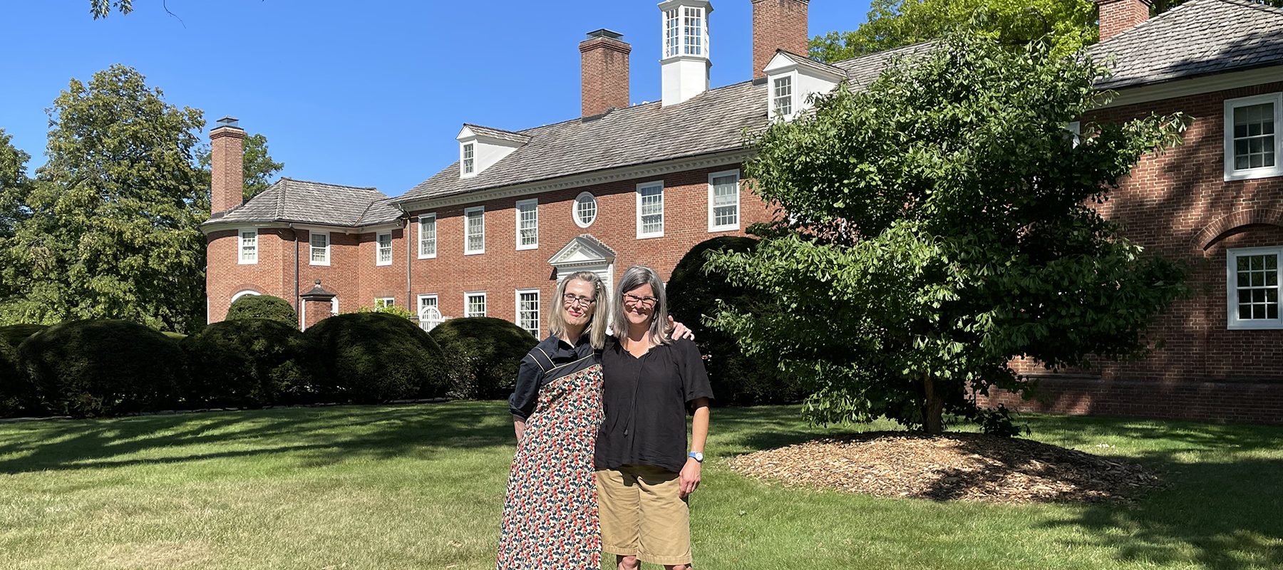 Two middle aged females stand in front of a large, colonial house.