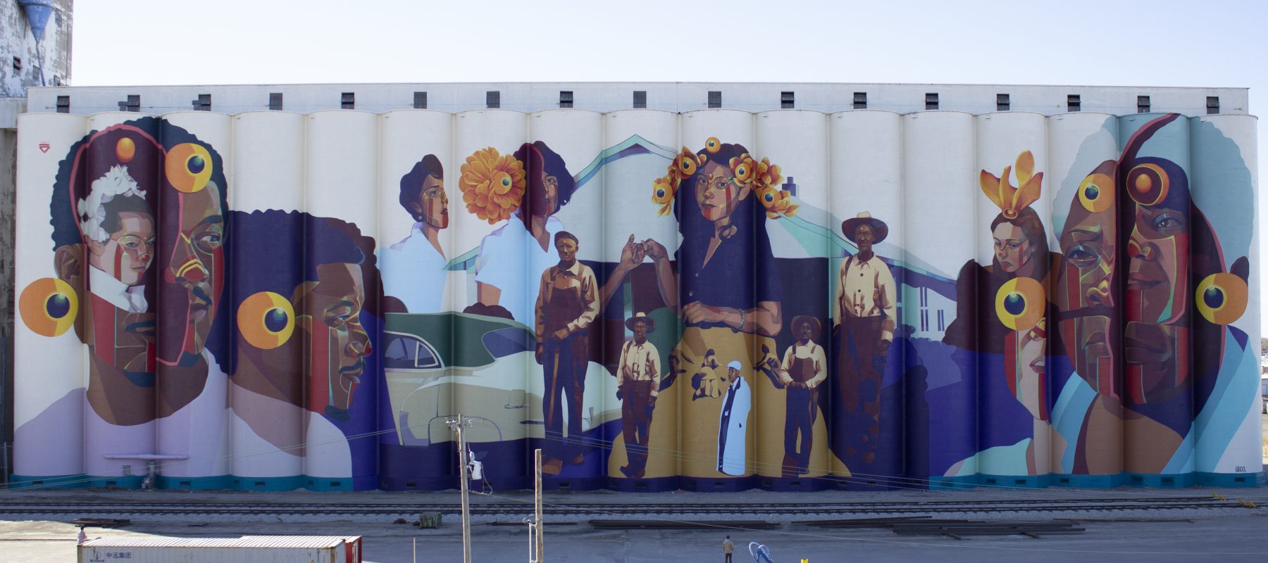 A huge mural is painted on the side of a grain elevator, featuring a variety of men and women of many backgrounds.