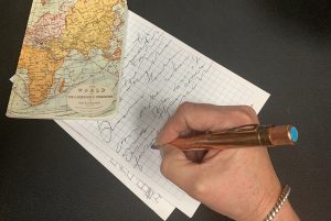 A woman's hand is holding a copper pen and writing on a pad of paper. A small notebook with a world map on it.