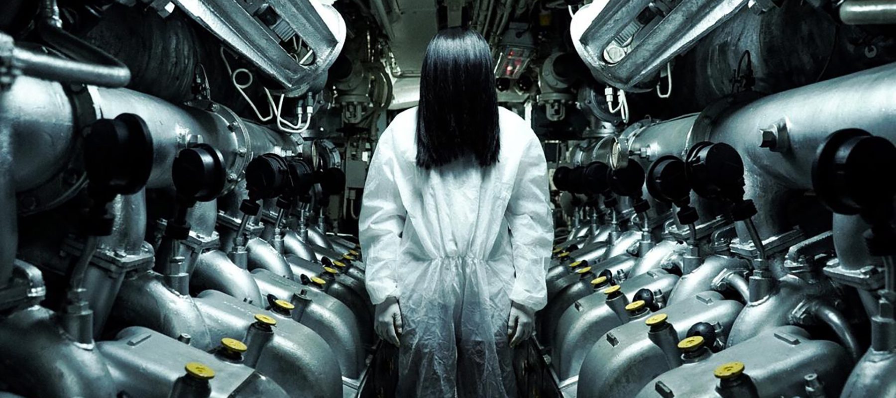 A person with long, black hair hiding their face stands between two rows of unidentified silver machines.