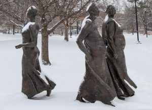 A statue of three ladies walking, one young, one middle aged and one old, covered in snow.