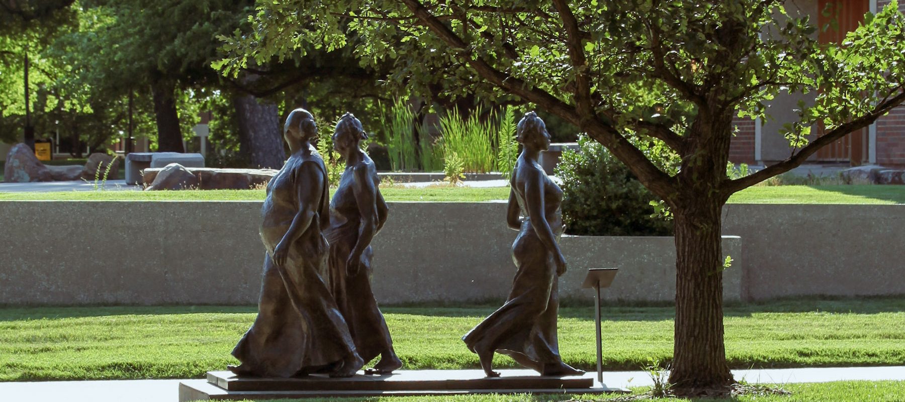 A bronze statue of three walking women is located under a shade tree.