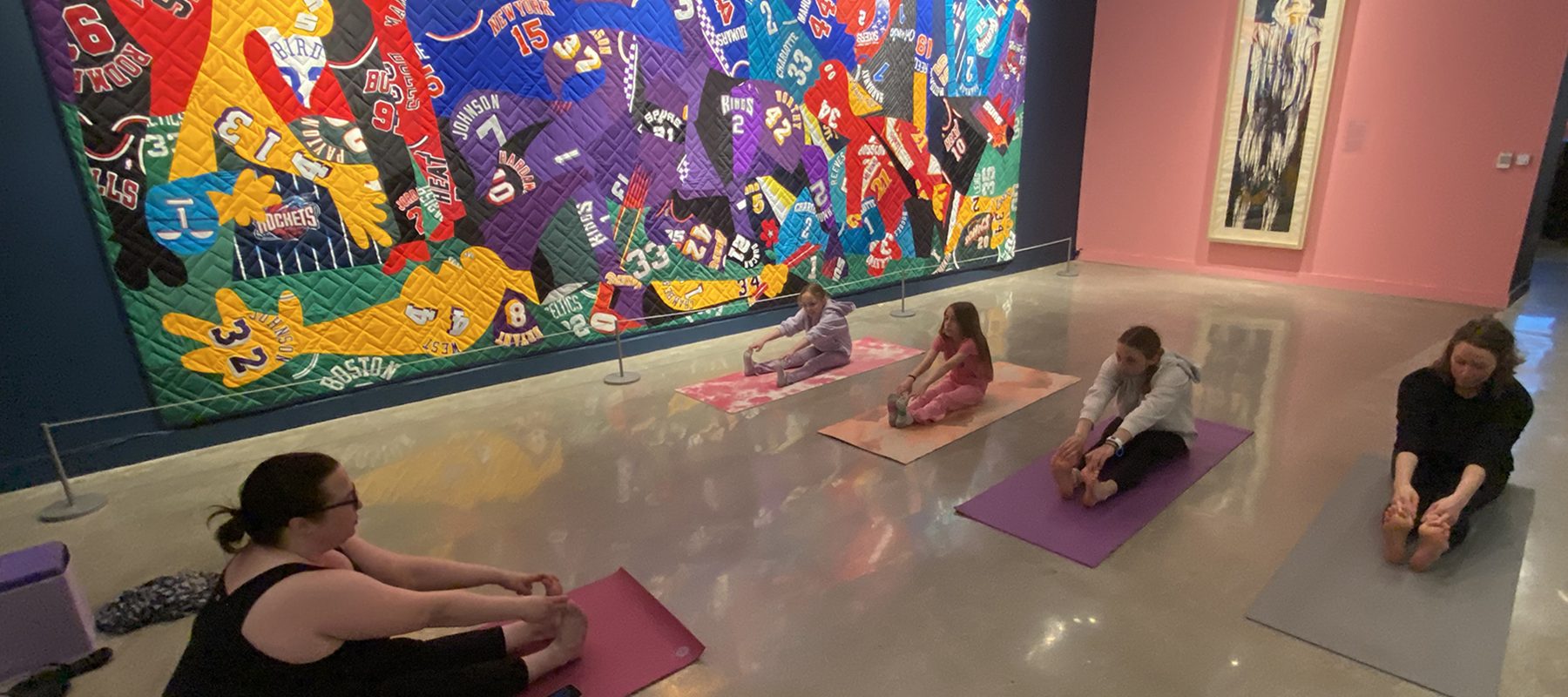 A yoga instructor leads three students in stretches in a gallery.