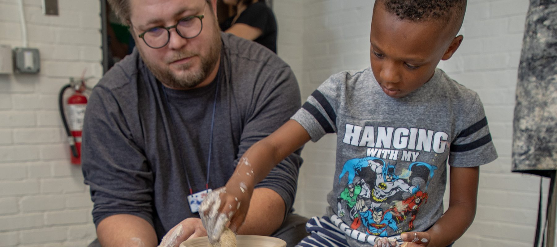 A man helps a boy throw a clay pot on a wheel.