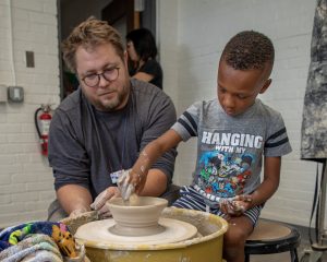 A man helps a boy throw a clay pot on a wheel.