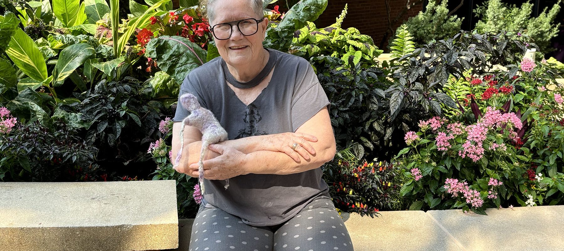 Artist Ernie Kind sits holding a felted figure in front of flowers.