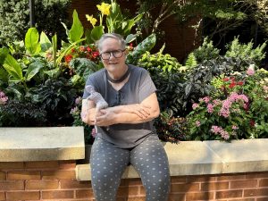 Artist Ernie Kind sits holding a felted figure in front of flowers.