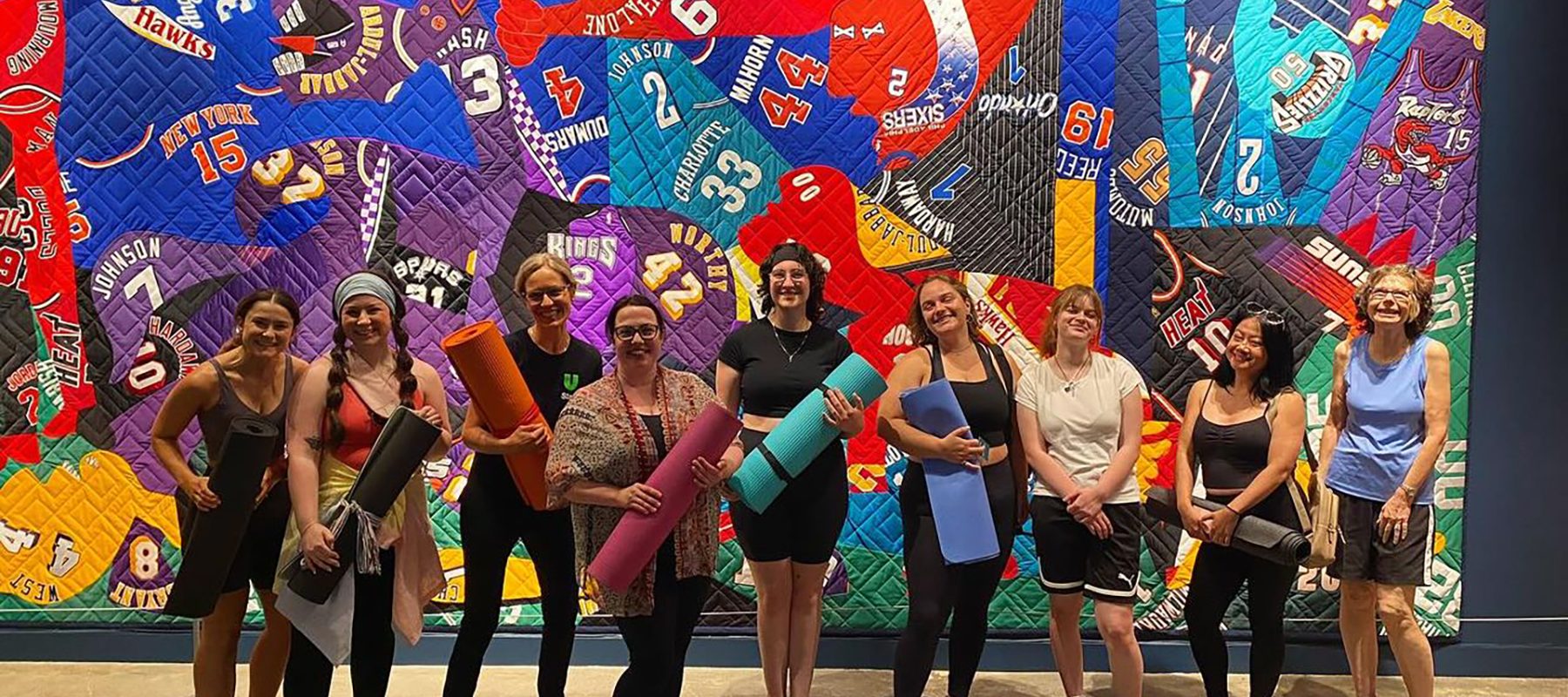 A line up of women holding yoga mats in a gallery. A large quilt hangs on the wall.