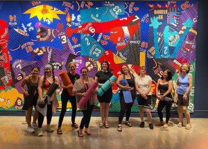 A line up of women holding yoga mats in a gallery. A large quilt hangs on the wall.