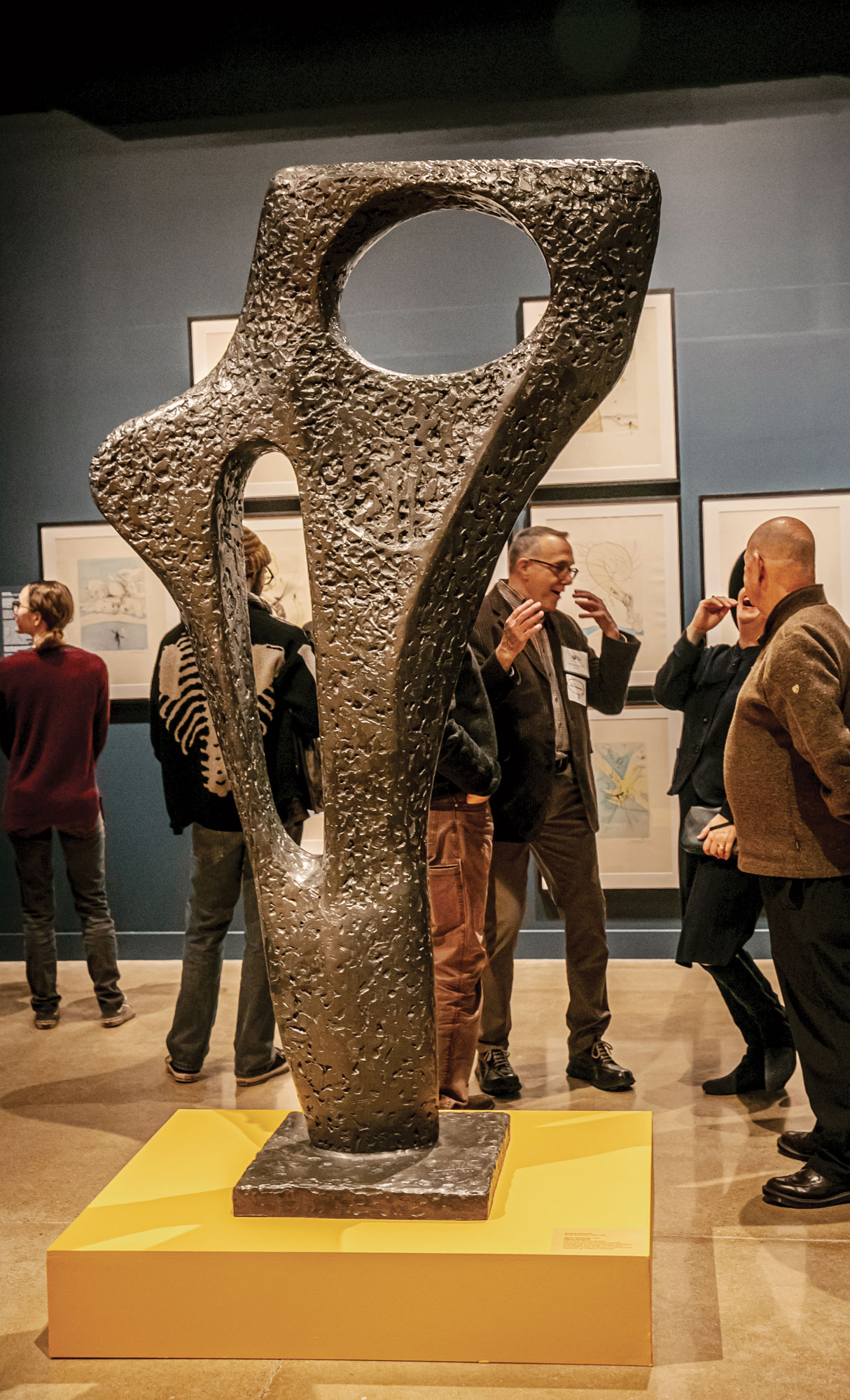 A man behind an abstract sculpture describes it with hand gestures to his friends