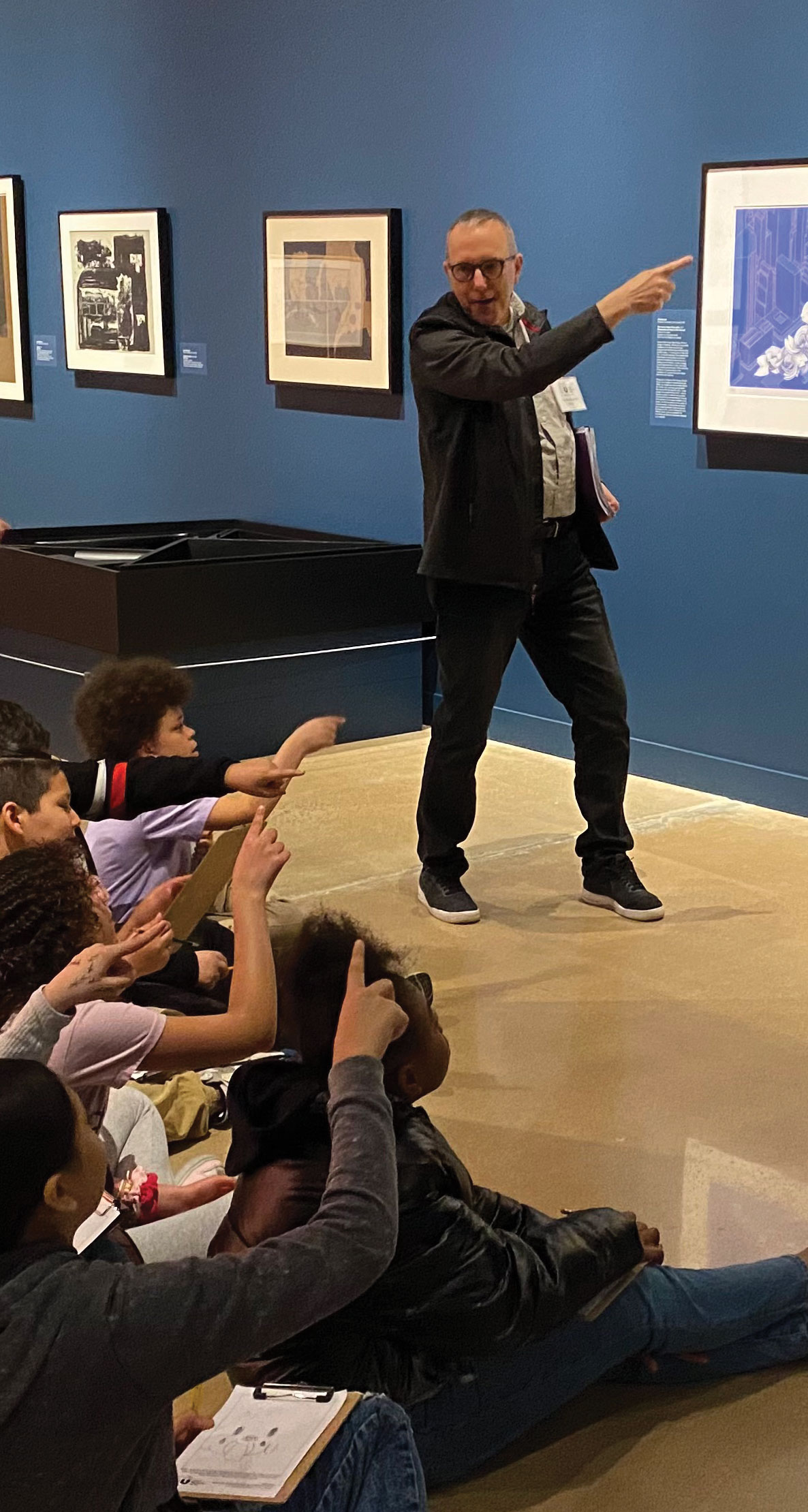 A man points at a painting while engaging students seated on the floor