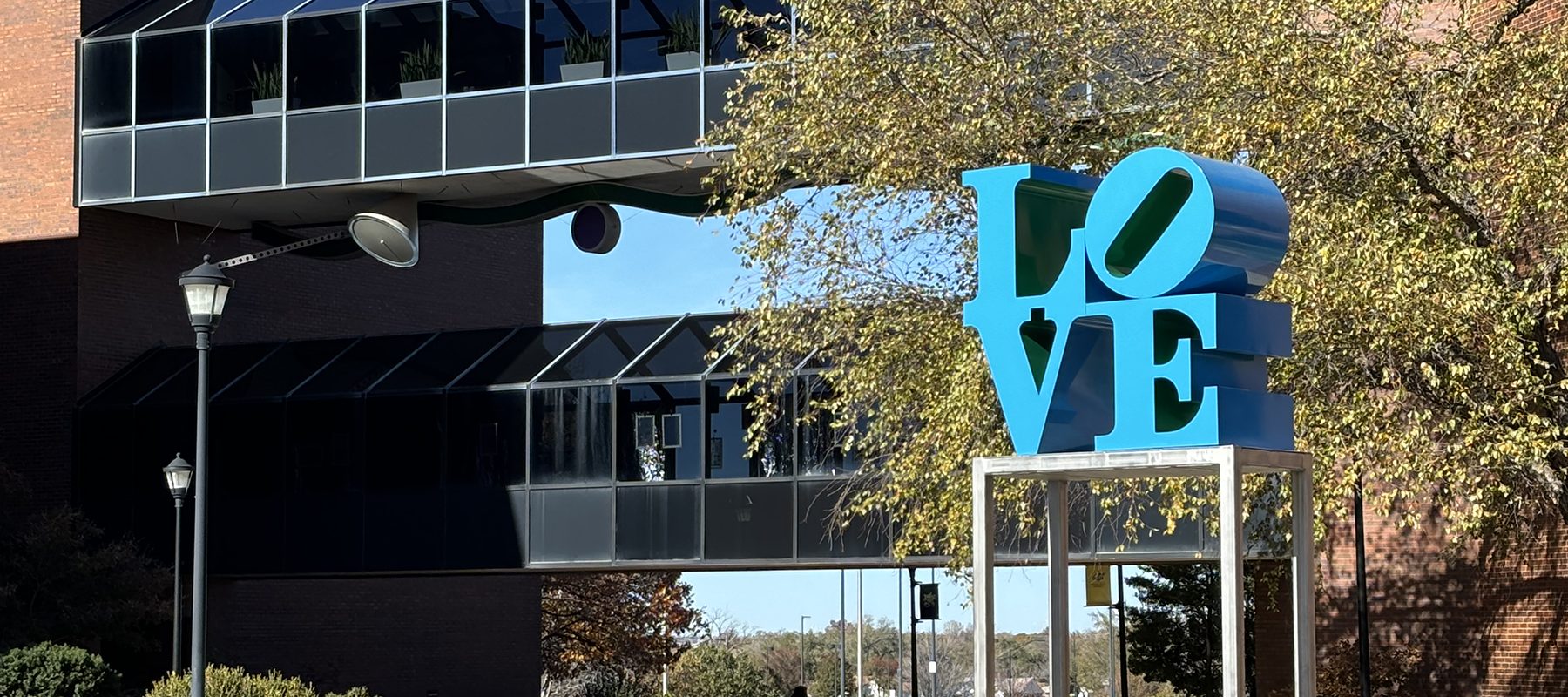 A large, blue LOVE sculpture sits atop a stand in front of the McKnight Art Center.