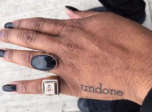 A hand with black nail polish, two rings and a tattoo that says, "undone."