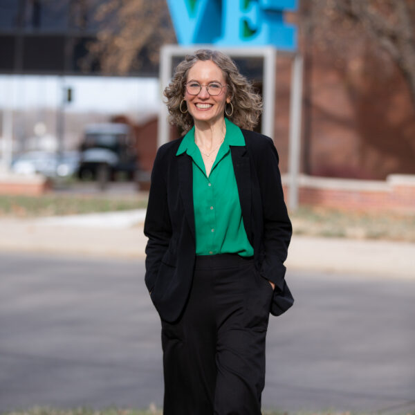 Blond woman in a black suit and green shirt smiles in front of the LOVE sculpture by Robert Indiana.
